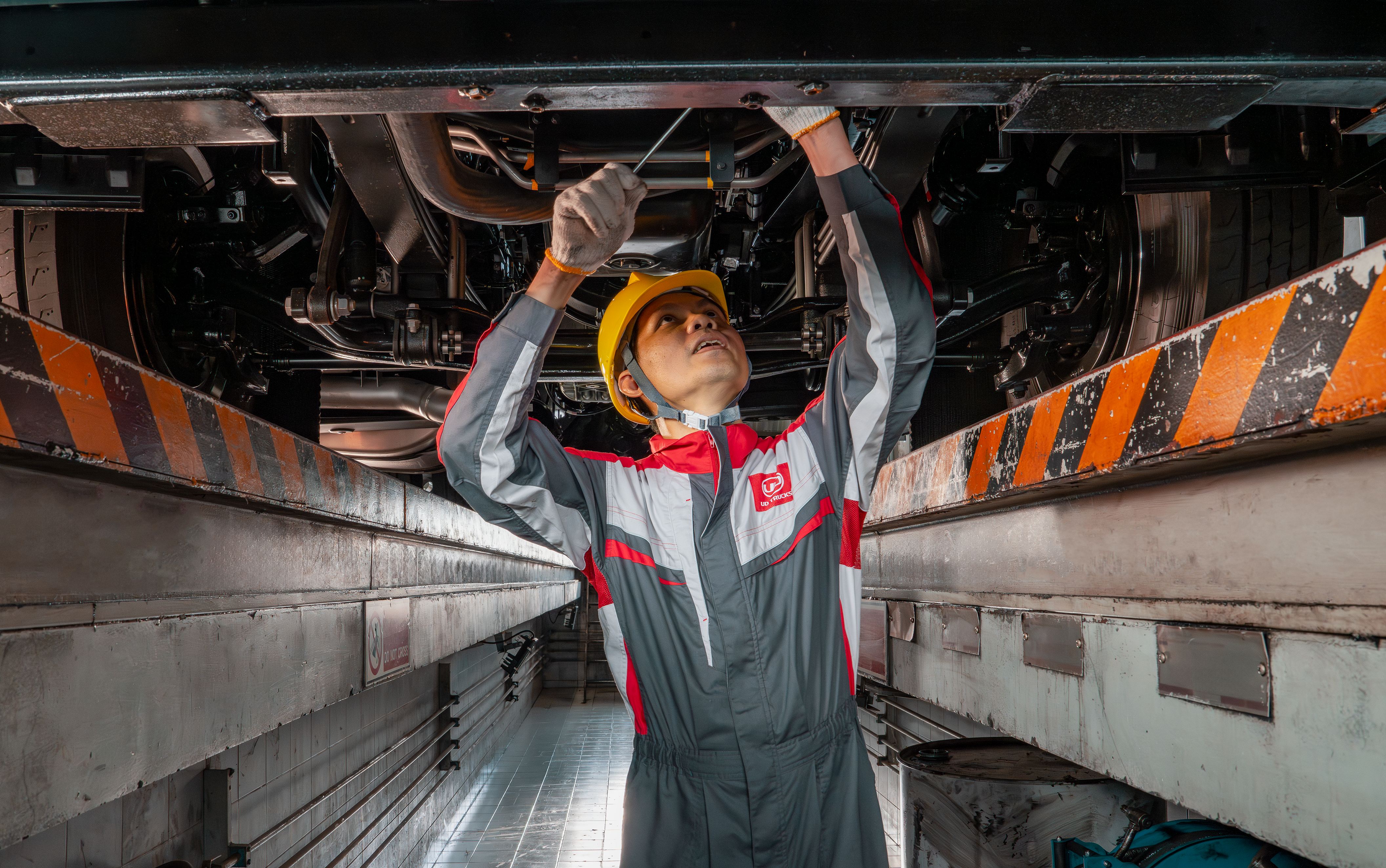 Aftersales Service Mechanic in coveralls and helmet working underneath a large vehicle in a service pit.