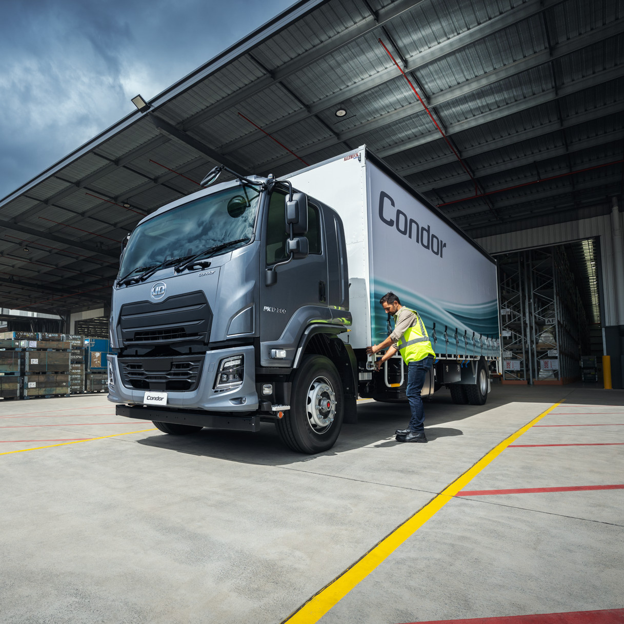 Truck parked at warehouse, worker in hi-vis vest beside it under cloudy sky.