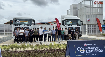 Grupo ng tao sa harap ng dalawang trak, may gusali at event banner sa likod.
